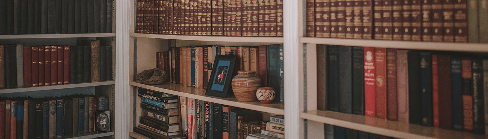 Corner of a library with white shelves full of vintage books, pottery and photos.