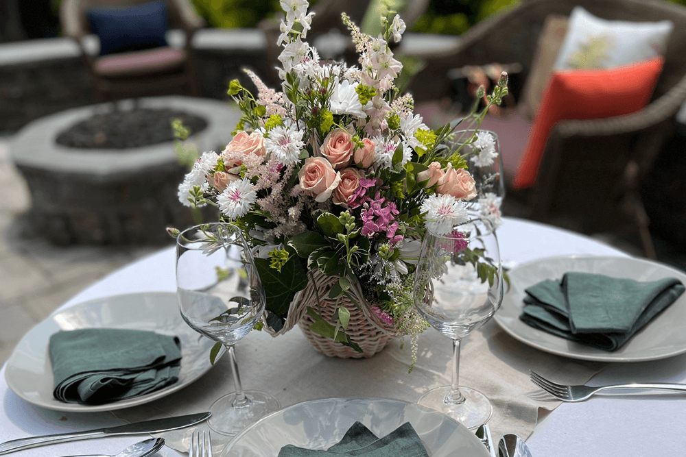 A small table set with a white cloth, green napkins, white plates, silver, wine glasses and a bouquet featuring peach roses, and other pink and white flowers with greenery.
