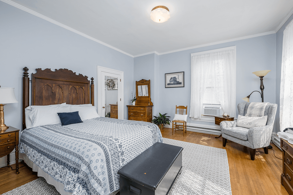 bedroom with wood headboard, blue and white striped cover, nightstand with light, wood cedar chest and padded chair, wood dresser with wood floors and white rug and blue walls
