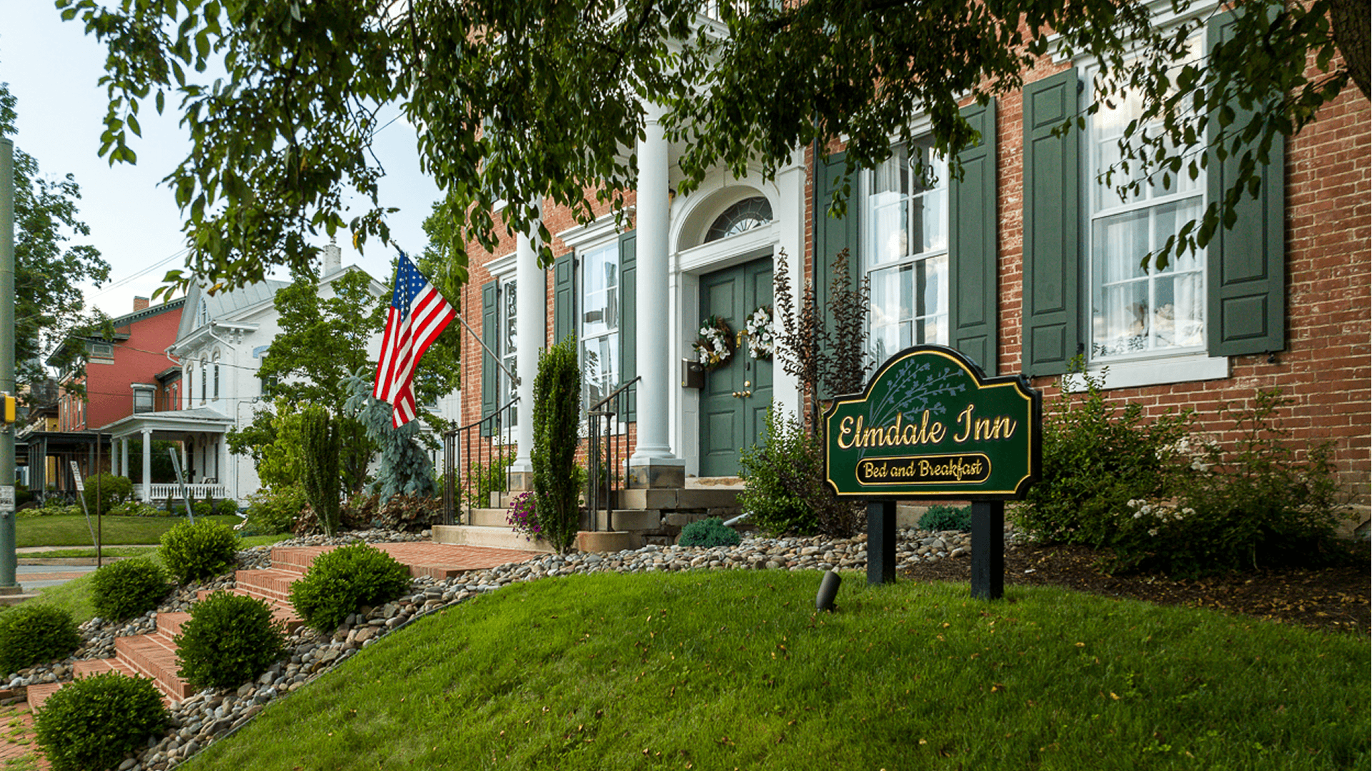 A large brick home with green shutters and door stands imposingly over a green sloped lawn. A wooden sign picked out in gold says Elmdale Inn Bed & Breakfast.