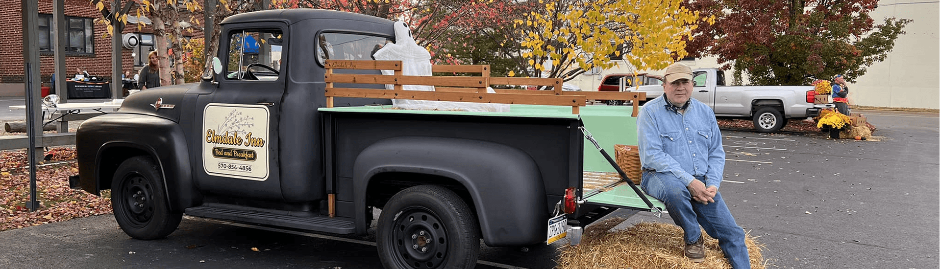 A man in denim sits on the tailgate of a vintage black truck with his foot resting on a hay bale. The truck is surrounded by fall foliage.