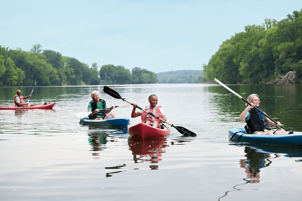 4 people in 4 kayaks, 2 red, 2 blue, on calm river with trees on both sides