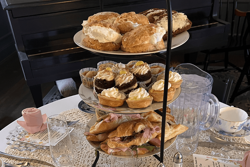 A table set for a Spring Tea with chinaware, silver, and a tiered tray with sandwiches, assorted pastries and cream puffs.
