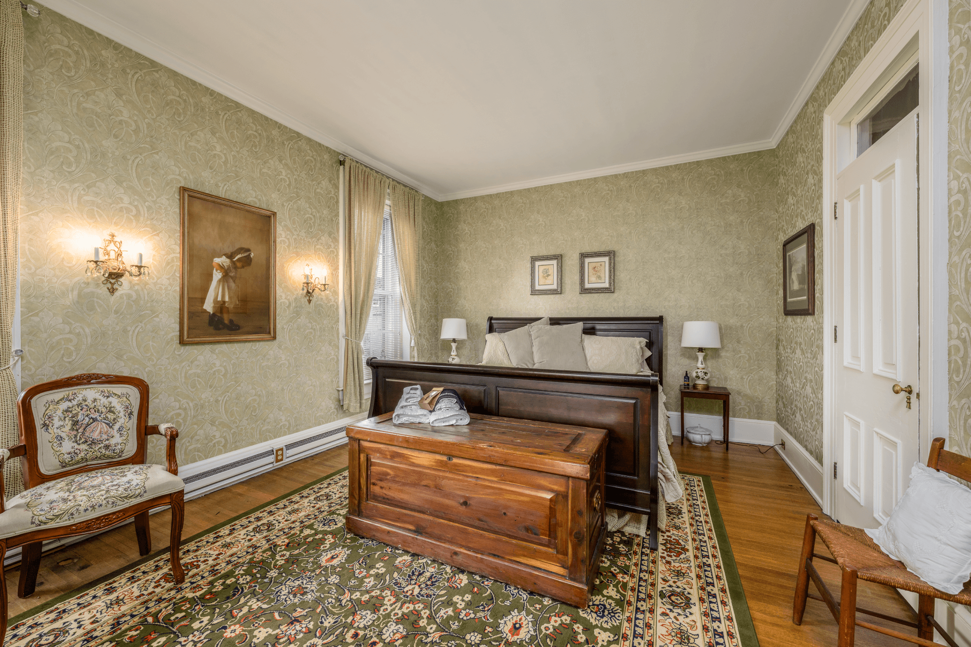 Room with king size sleigh bed, wooden chest & green wallpaper with artwork on the walls