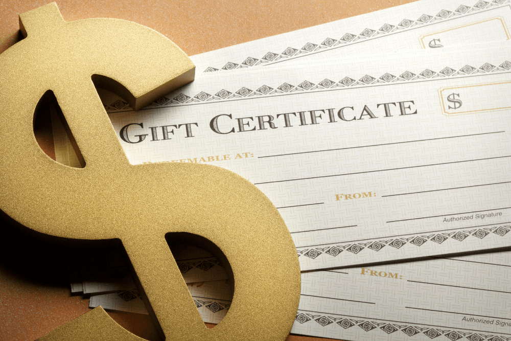 Multiple blank paper gift certificates spread out on a wood table with a gold Dollar sign paper weight holding them down