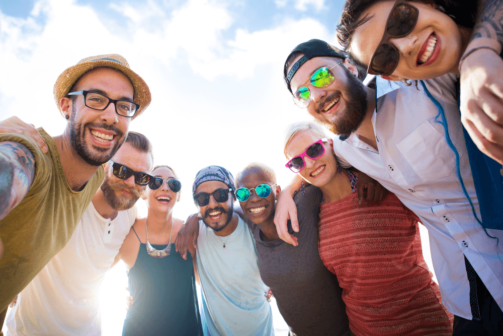 Group of males and females of different races, with an assortment of hats and sunglasses standing in a semicircle with arms interlocked with blue sky and clouds behind them