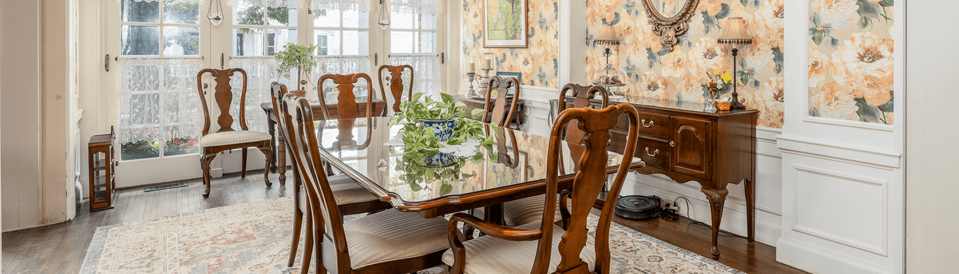 Dining room with floral wallpaper, wall mirror, buffet, horizontal table with matching chairs windows streaming in lights
